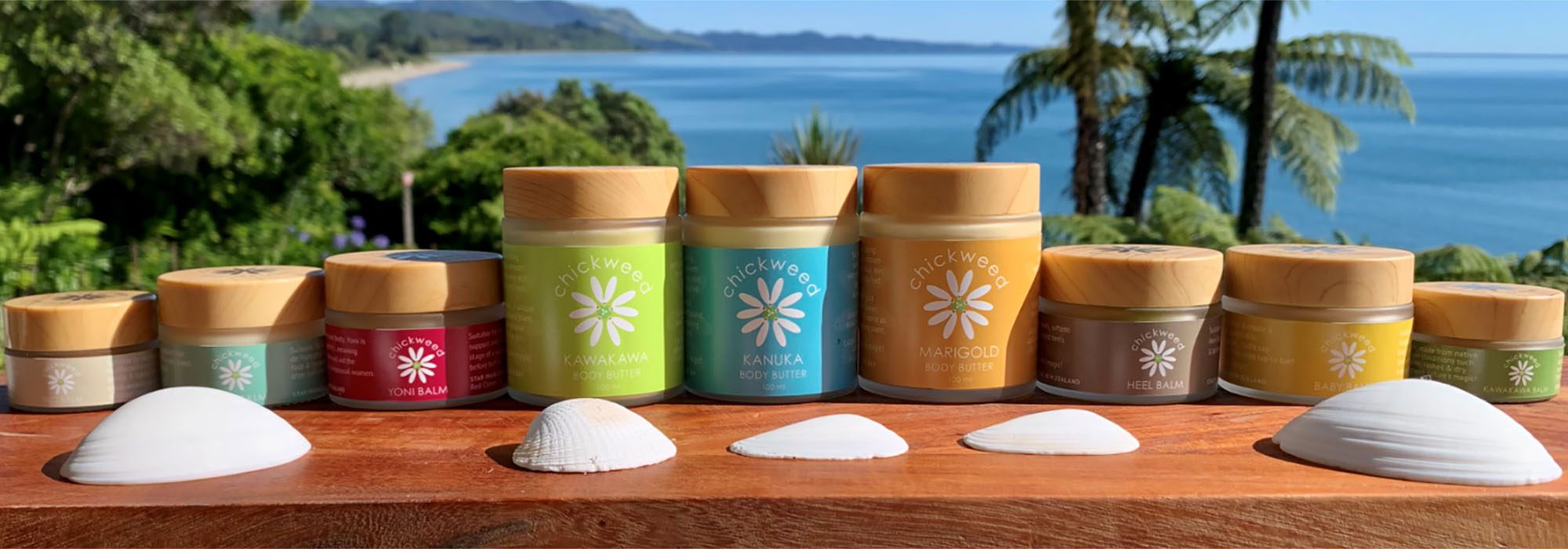 Image of various coloured pots containing body butters an balms, with a backdrop of a sunny Golden Bay beach and palm trees